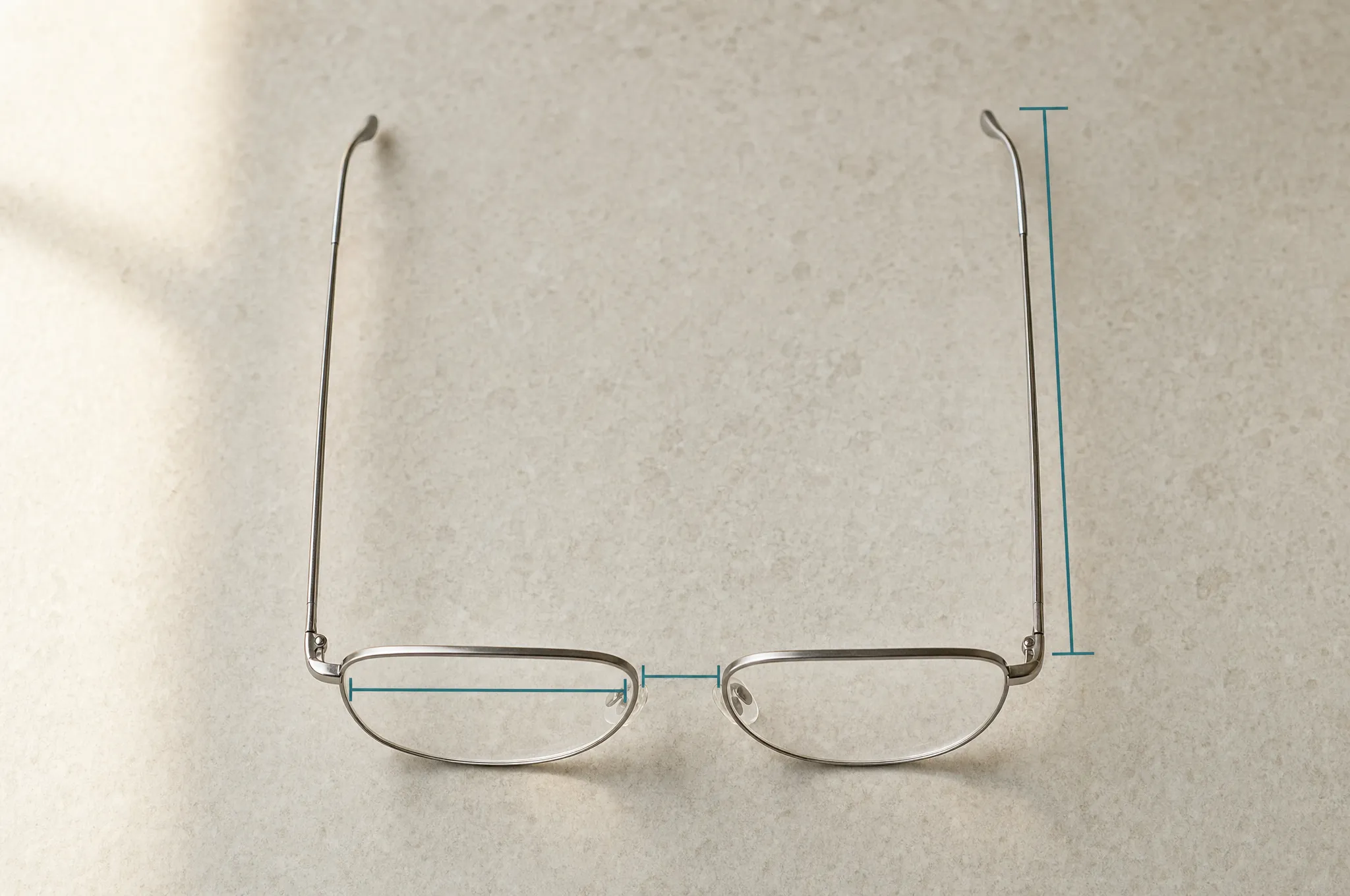 Glasses lying open on stone, seen from above, with three drawn spans marking one lens width, the bridge gap and one temple arm